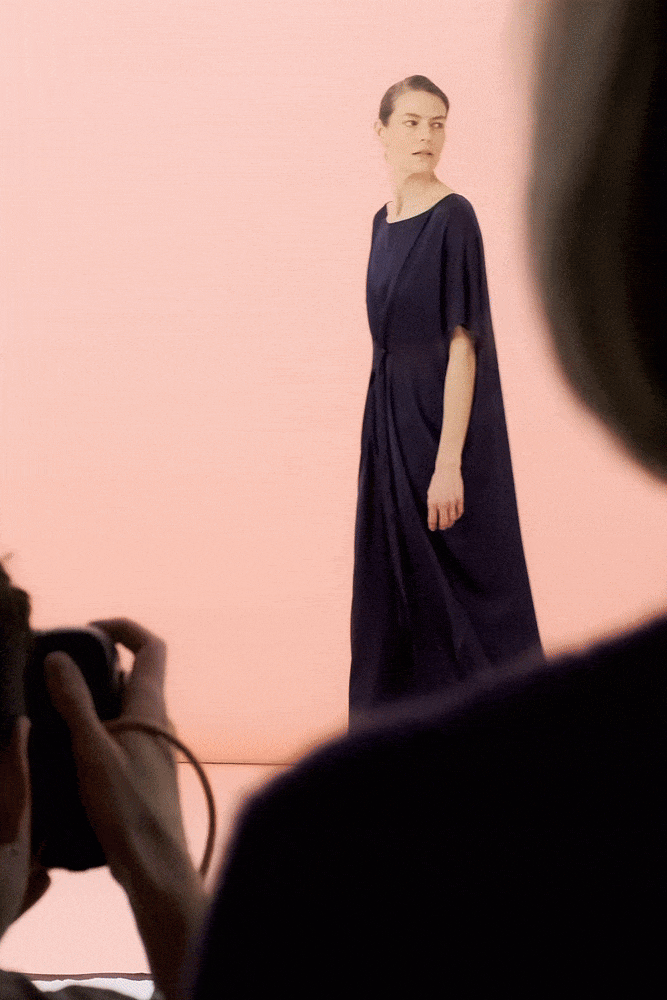 Model walking in a navy jewel silk satin caftan dress with a belted waist and fluid drape, on a soft blush pink studio backdrop during a photoshoot.